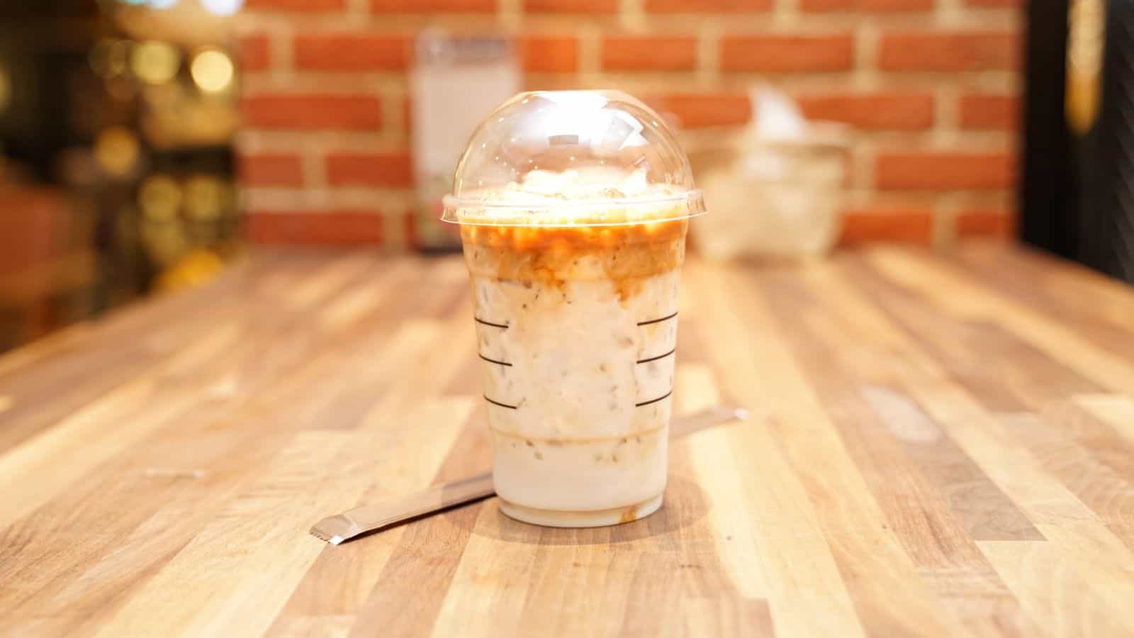 A plastic cup filled with iced coffee topped with caramel sauce and ice, sitting on a wooden table. A dome lid covers the cup, and a plastic straw is placed next to it. A blurred brick wall is in the background.