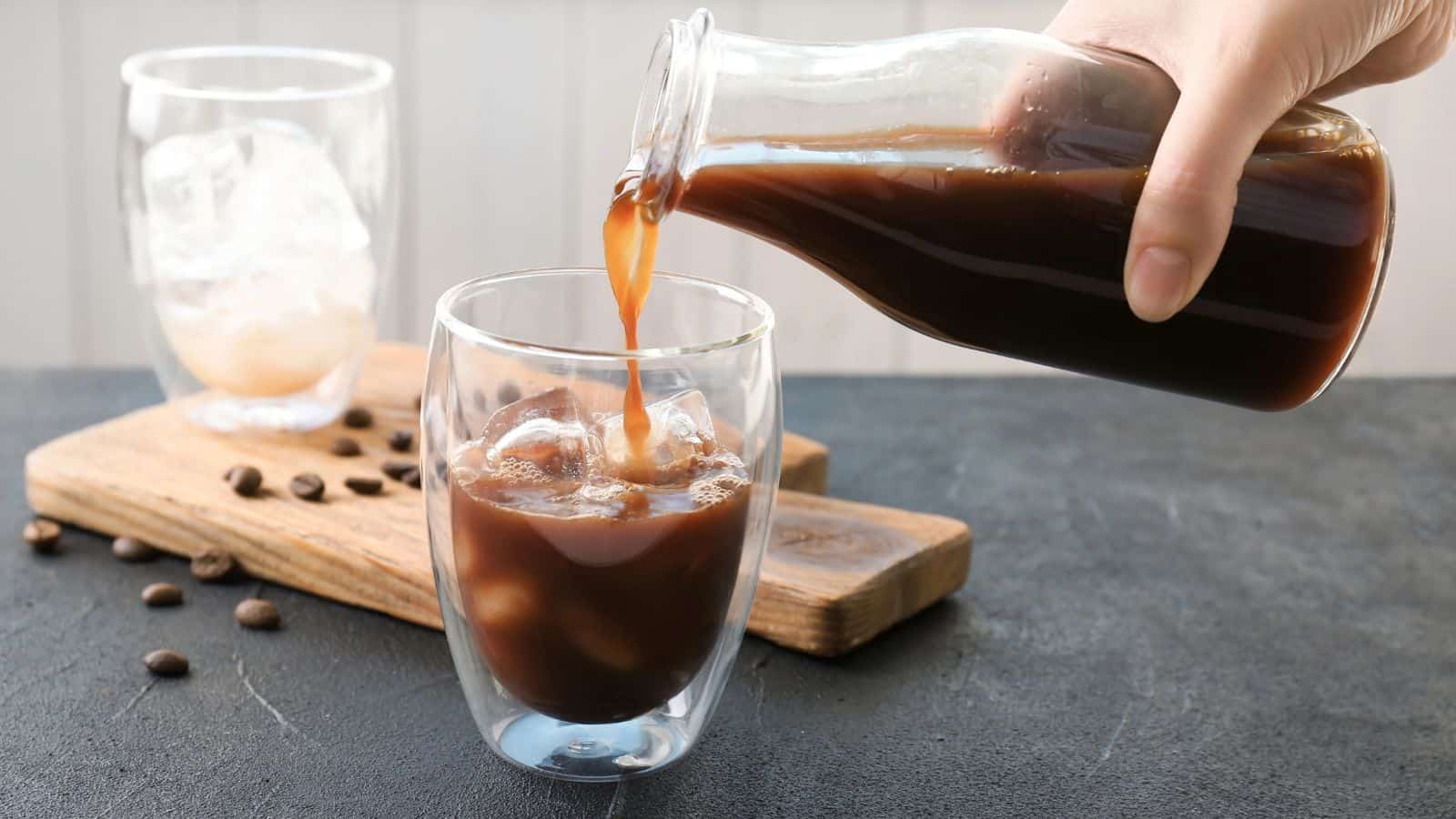 A hand pours iced coffee from a glass bottle into a double-walled glass filled with ice. Another glass with ice and a few coffee beans on a wooden board are in the background.
