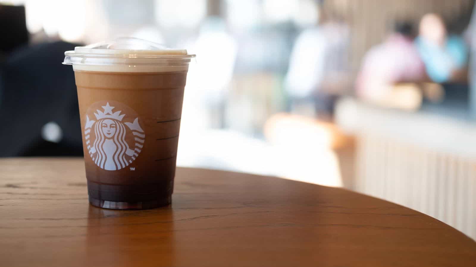A plastic cup of iced coffee with cream and a lid, featuring the Starbucks logo, sits on a wooden table in a blurred café setting.