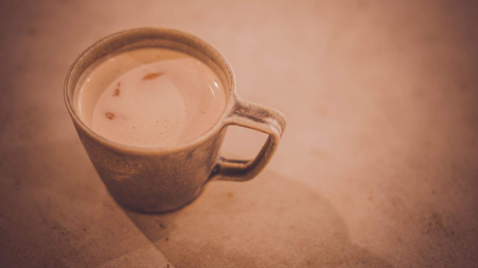 A ceramic mug filled with light brown coffee or tea sits on a light beige surface. The mug has a visible handle and some reflections are seen on the drink’s surface.