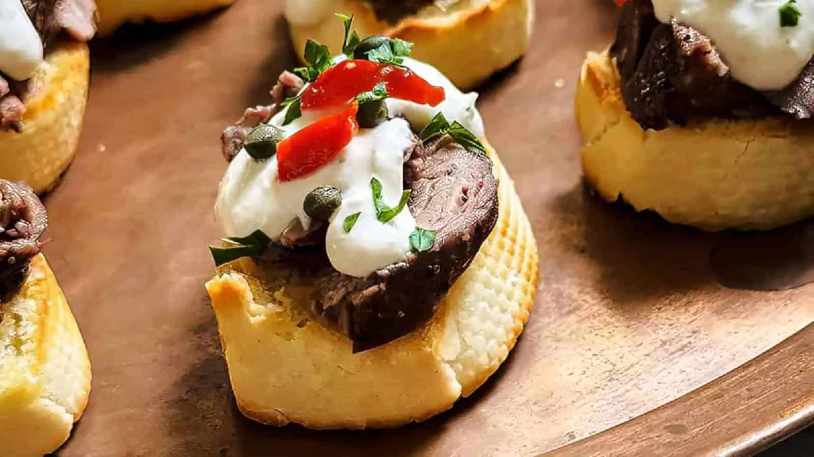 Open-faced sandwiches with sliced roast beef, creamy sauce, red peppers, and capers on a parchment-lined surface.