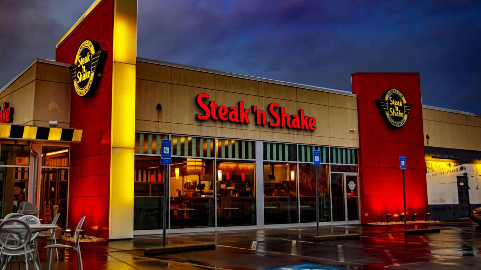 The exterior of a Steak 'n Shake restaurant at dusk, with illuminated red and yellow signage, large windows, and a wet parking lot with visible handicapped parking spaces in front.