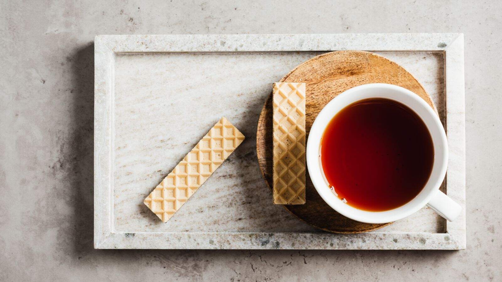 A white cup filled with black tea sits on a round wooden coaster. Two rectangular wafer biscuits lie next to the cup on a rectangular marble tray. The background is a smooth, light gray surface.