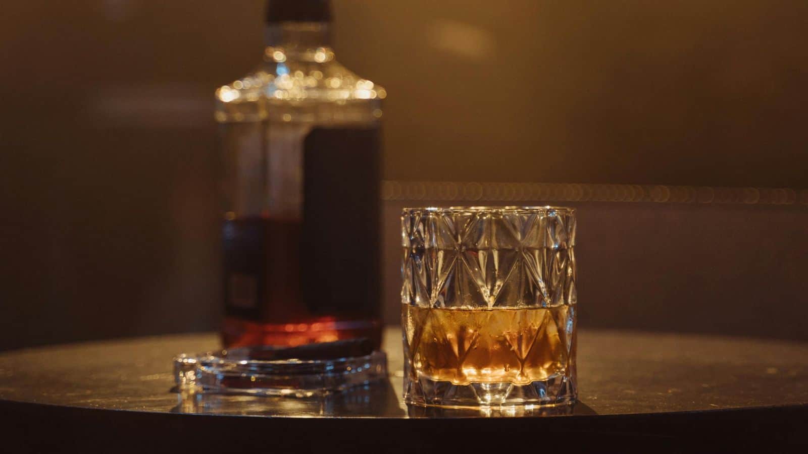 A glass of whiskey sits on a round table next to a partially filled whiskey bottle and an ashtray. The background is blurred and dimly lit.