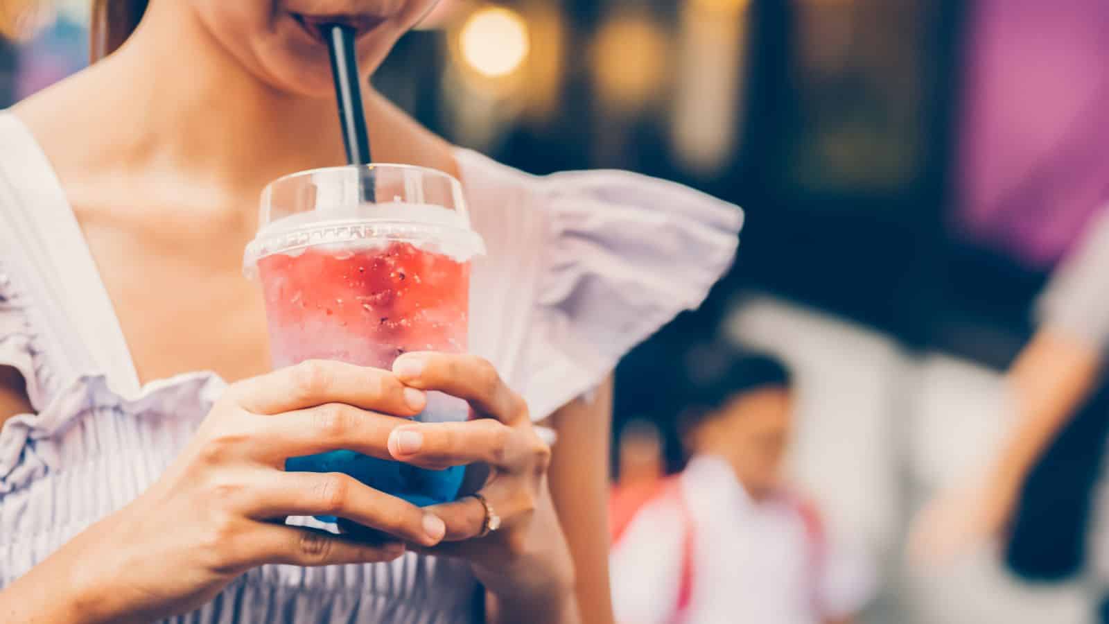 A person drinks from a clear cup with a black straw, containing a layered red and blue iced beverage. The person is wearing a white, ruffled top. The background is blurred, with indistinct figures and lighting.