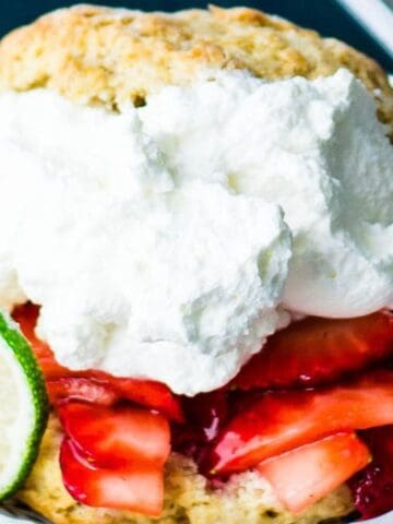 A dessert with sliced strawberries and whipped cream layered on a biscuit is pictured. A lime slice is garnishing the dish, which is served in a white bowl on a dark surface. A fork is partially visible to the right.