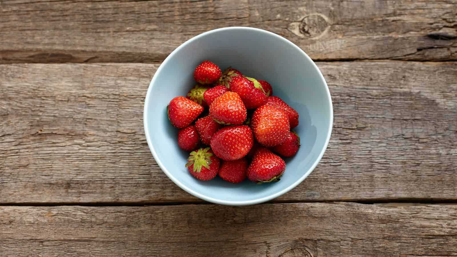 A light blue bowl filled with red strawberries sits on a rustic wooden surface.