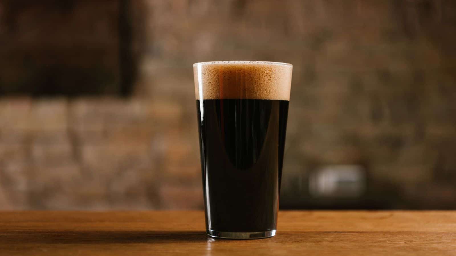 A pint glass filled with dark stout beer topped with a thick, creamy foam head sits on a wooden surface, with a blurred brown brick wall in the background.