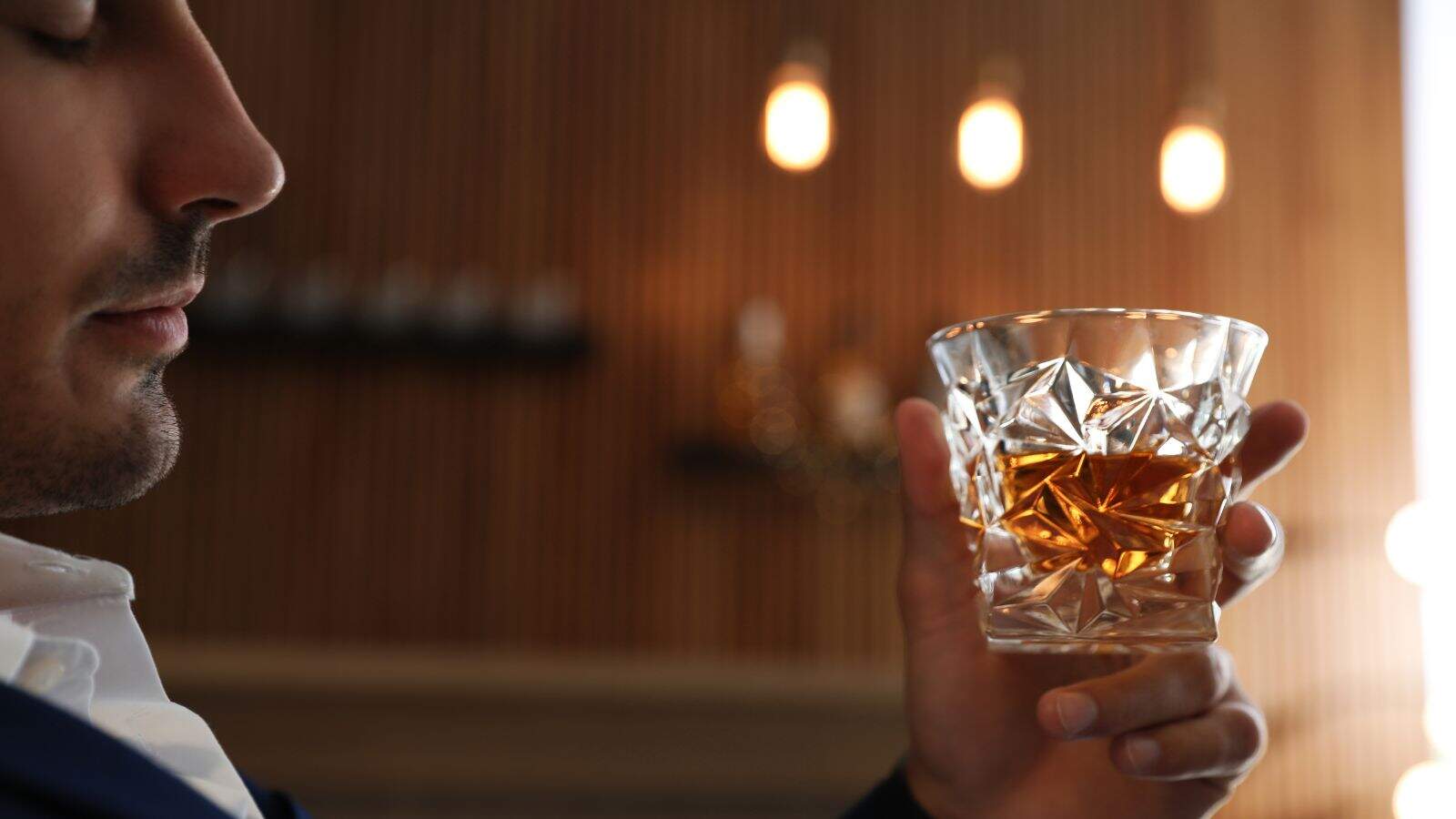 A person holds a faceted glass filled with amber liquid, possibly whiskey, near their face with eyes closed. The background is softly lit with hanging lights and wooden paneling.