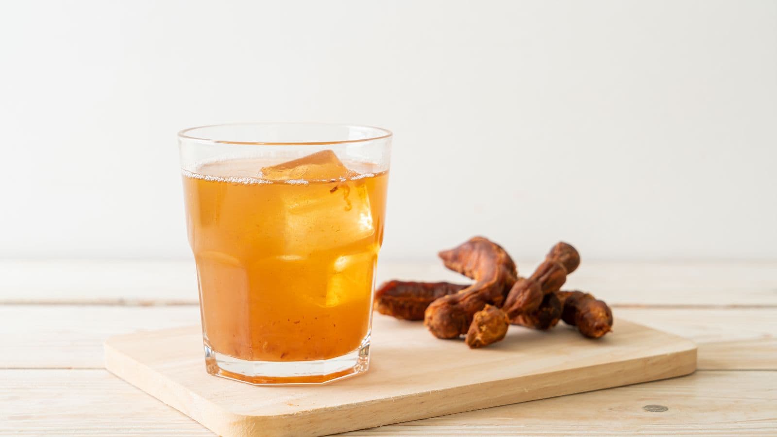 A glass of iced juice with tamarind pods on a wooden board. The background is plain white, and the board is placed on a light wood surface.