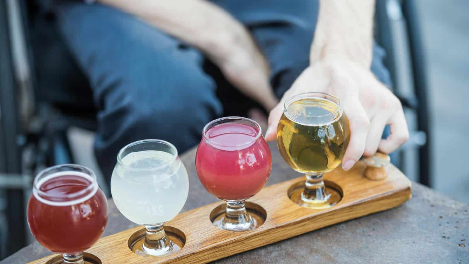 A person in dark clothing holds a wooden paddle with four small glasses filled with different colored drinks&mdash;red, pale yellow, pink, and golden&mdash;arranged in a row.