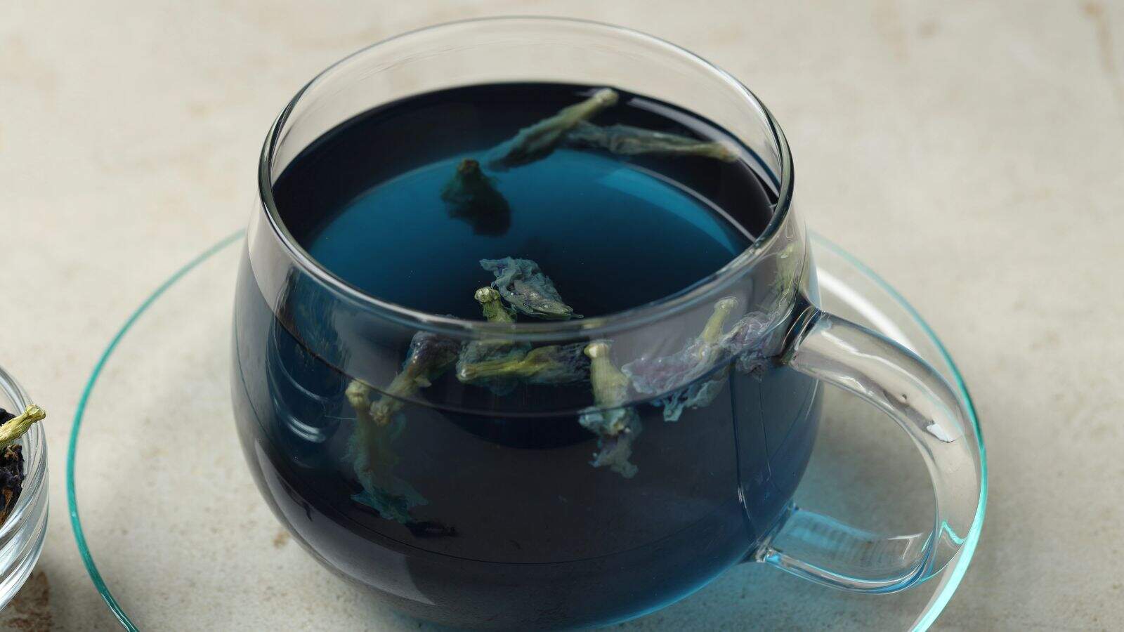 A clear glass cup sits on a glass saucer, filled with deep blue butterfly pea flower tea. Several dried flower petals can be seen floating on the surface. The table beneath is a light beige color.