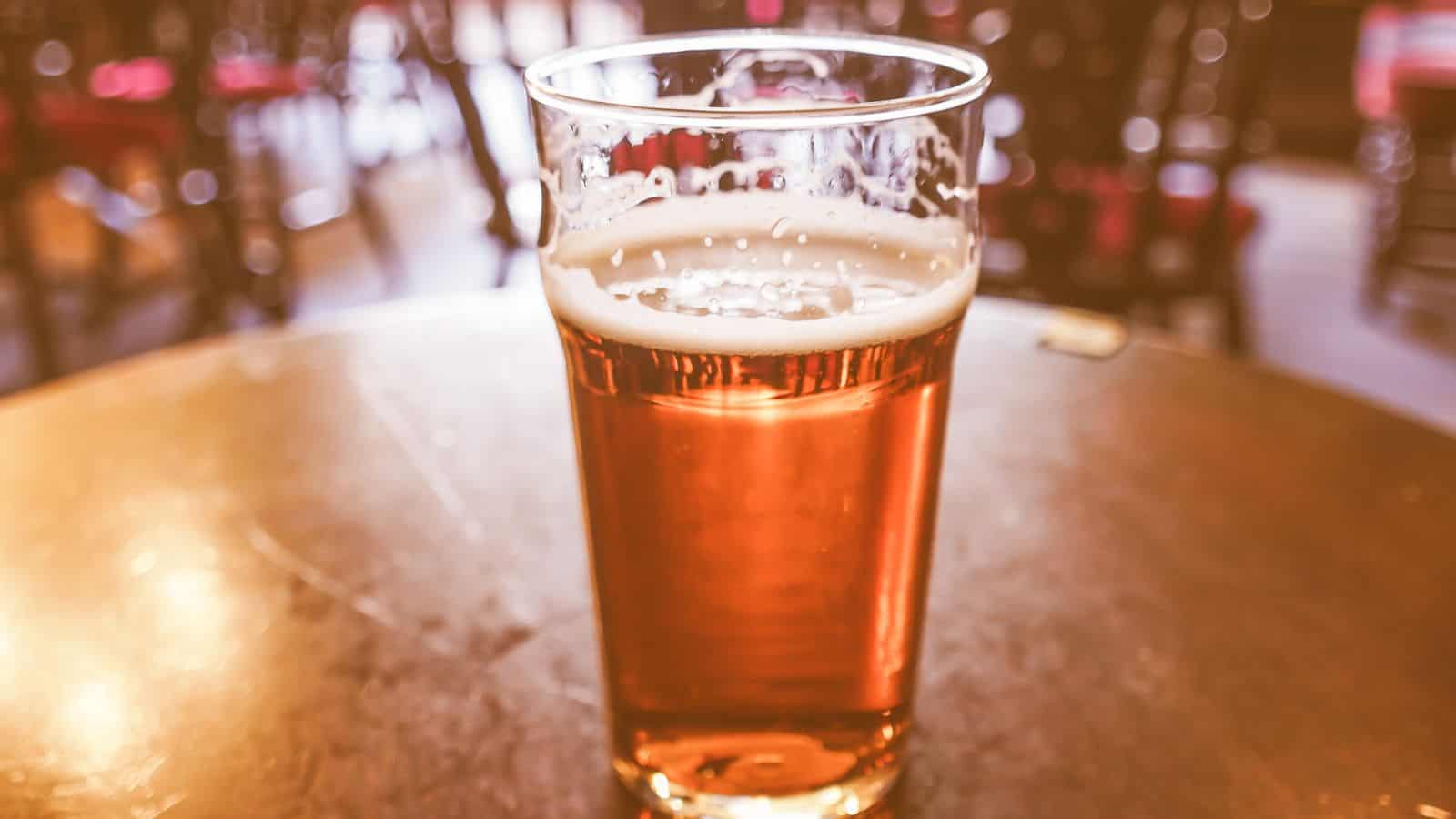 A glass of amber-colored beer sits on a wooden table in a pub setting. The glass is half-full, with a small frothy head on top. Out of focus in the background are chairs and tables, suggesting a relaxed atmosphere.