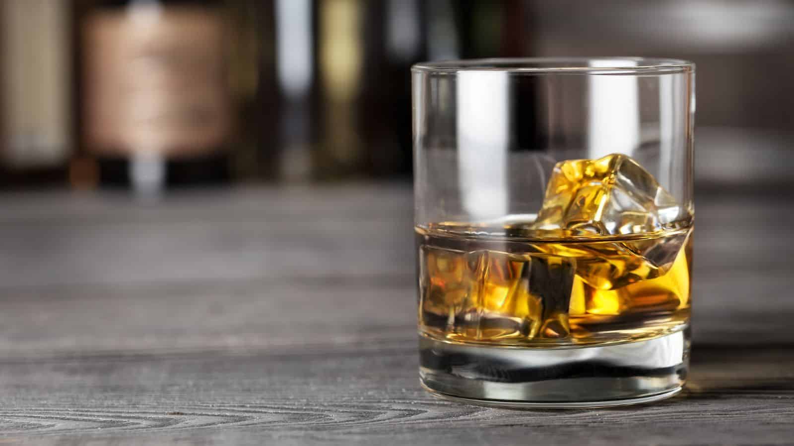 A glass of whiskey with ice cubes sits on a wooden surface, with blurred bottles and a neutral background in the distance.