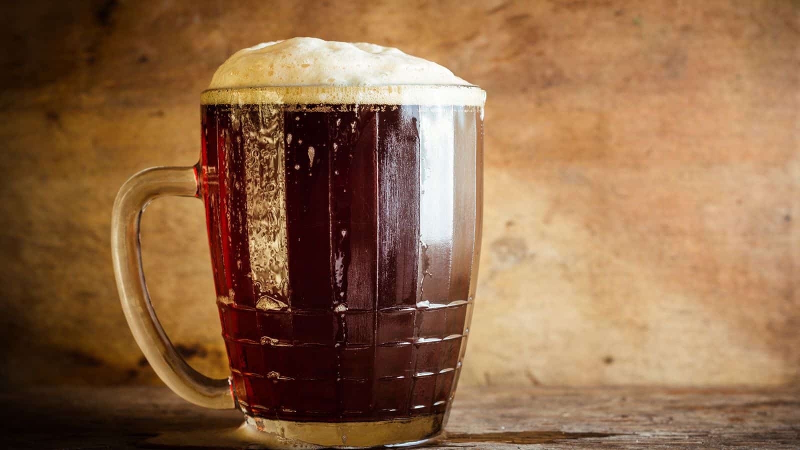 A glass mug filled with dark beer, topped with a frothy head. The mug is on a wooden surface with a textured, brown wooden background. Some condensation is visible on the sides of the glass.