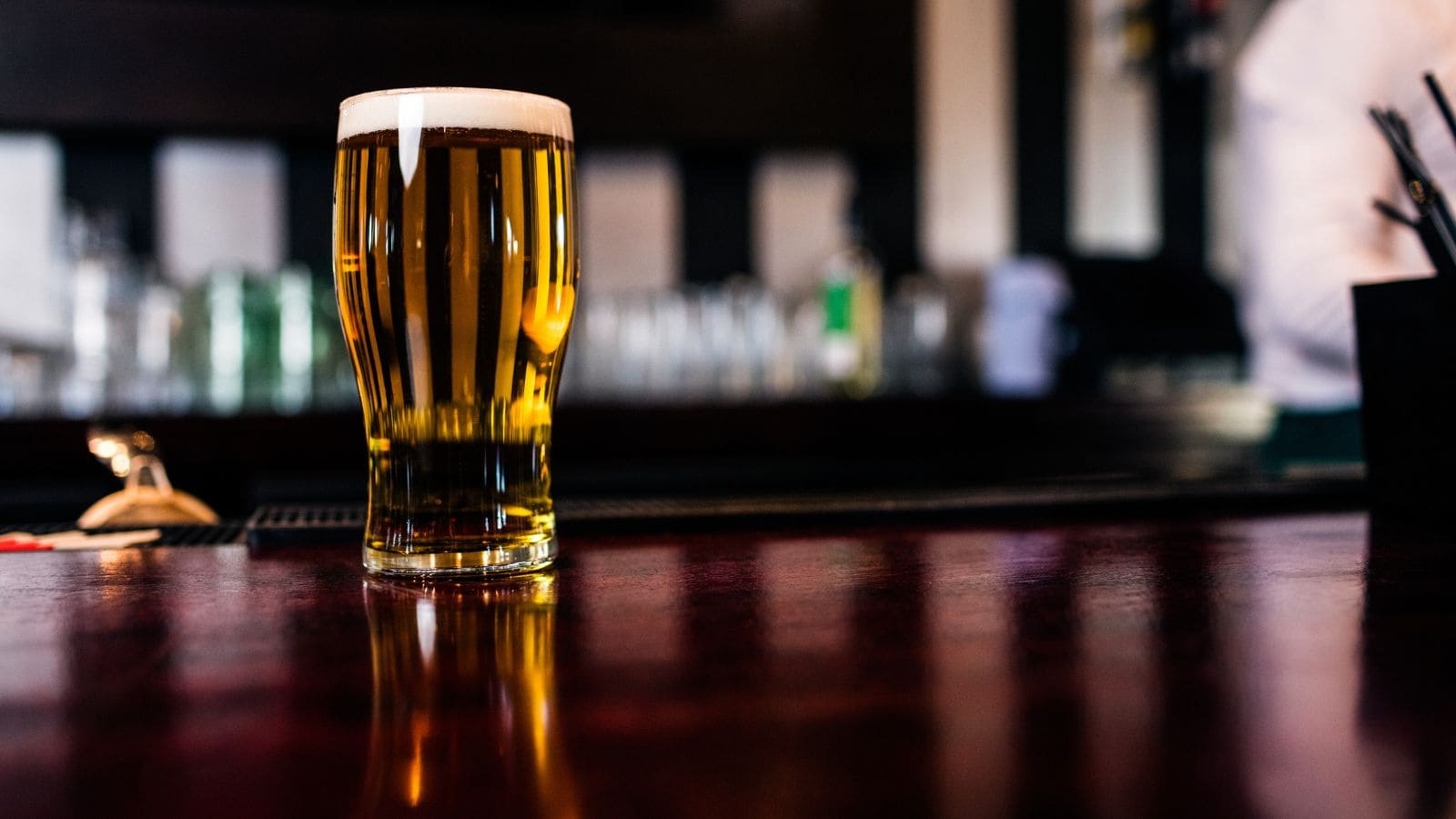 A pint glass filled with light beer sits on a polished wooden bar counter, with a blurred background showing bar equipment and part of a person in white.