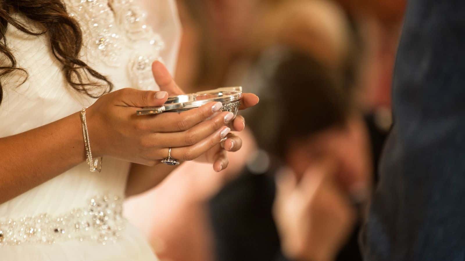 A person in a white, beaded dress holds a small, round dish with both hands. The focus is on the hands and dish, with blurred people in the background.