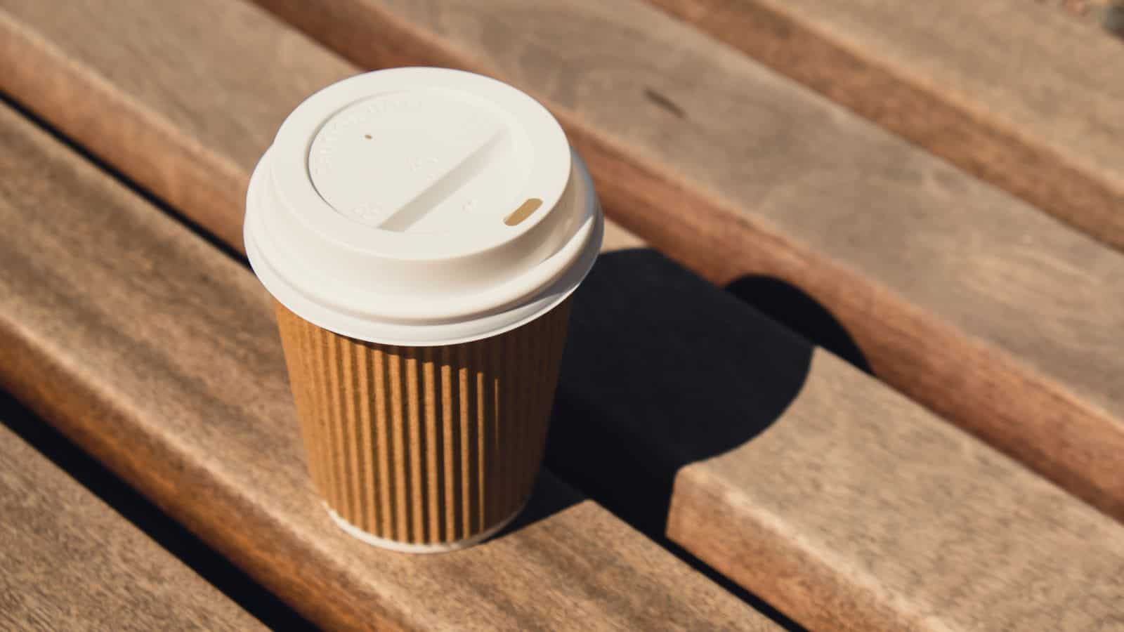 A brown disposable coffee cup with a white plastic lid sits on a wooden bench under sunlight. The cup casts a shadow on the bench surface.