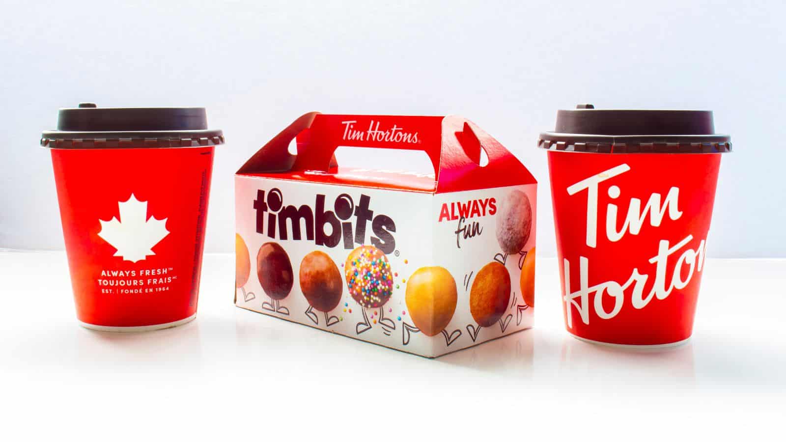 Two red Tim Hortons coffee cups with brown lids flank a Tim Hortons Timbits box. The box features images of various donut holes and the words "timbits" and "ALWAYS fun." The cups display the Tim Hortons logo and a maple leaf.