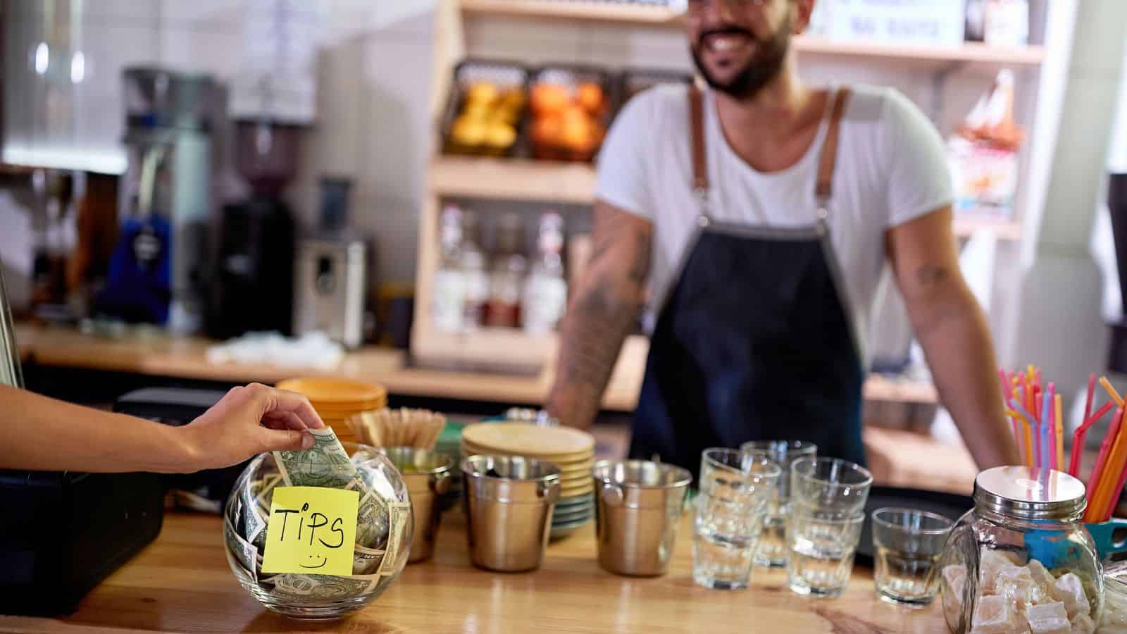 A person places money into a glass tip jar labeled "TIPS" on a cafe counter. The barista, wearing a white shirt and dark apron, stands behind the counter, smiling. Glasses and cups are neatly arranged on the wooden countertop.