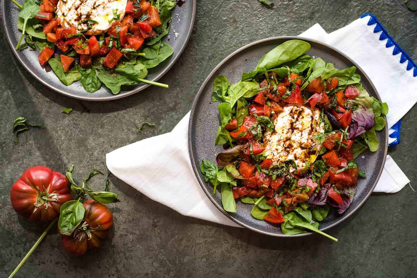 A salad with spinach, tomatoes, and a scoop of burrata drizzled with balsamic glaze is served on a dark plate. A similar salad sits nearby, with heirloom tomatoes and basil leaves to the side on a textured green surface. A white napkin lies beneath the plates.