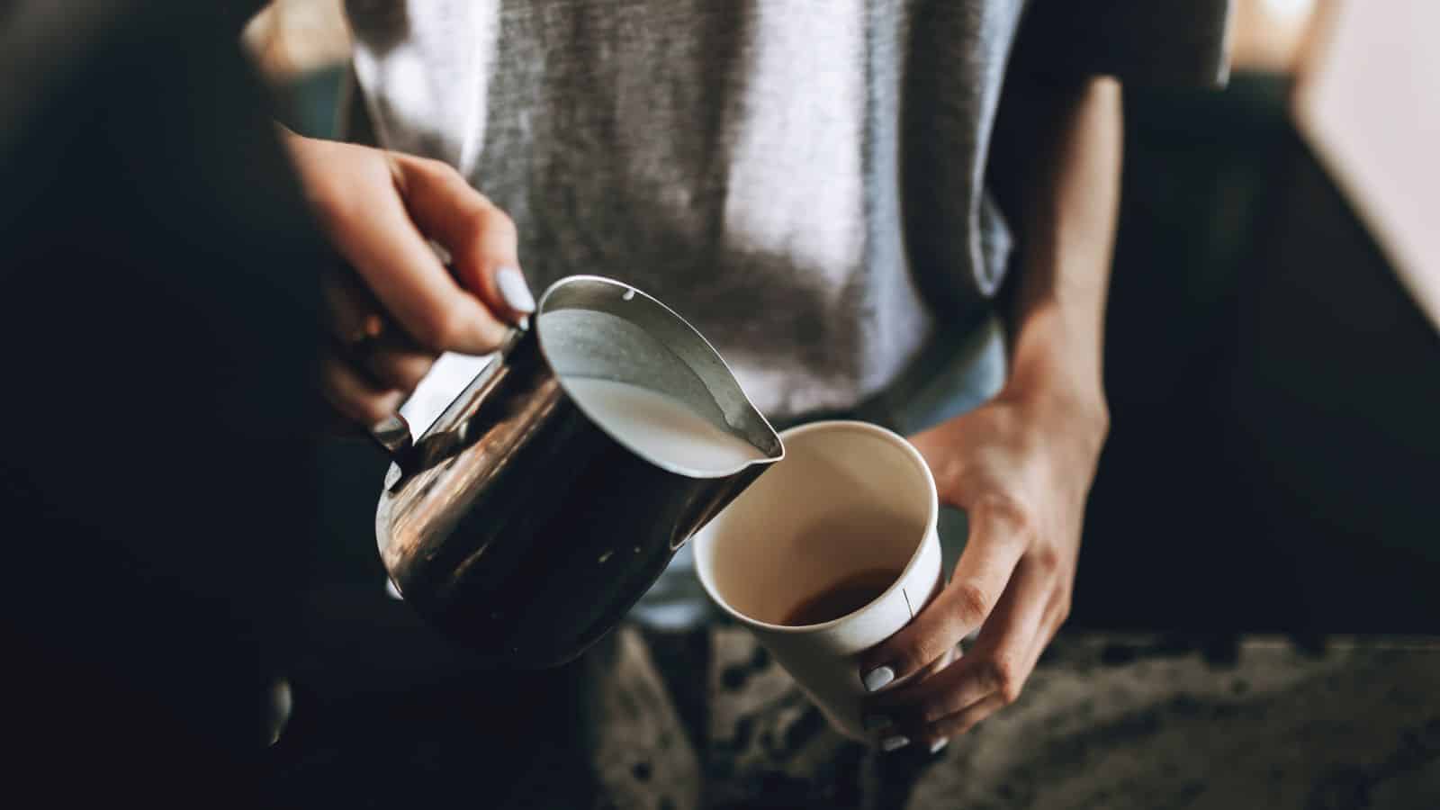 A person is pouring milk from a metallic pitcher into a paper cup filled with coffee. The person is wearing a gray top, and the countertop appears to be made of stone.