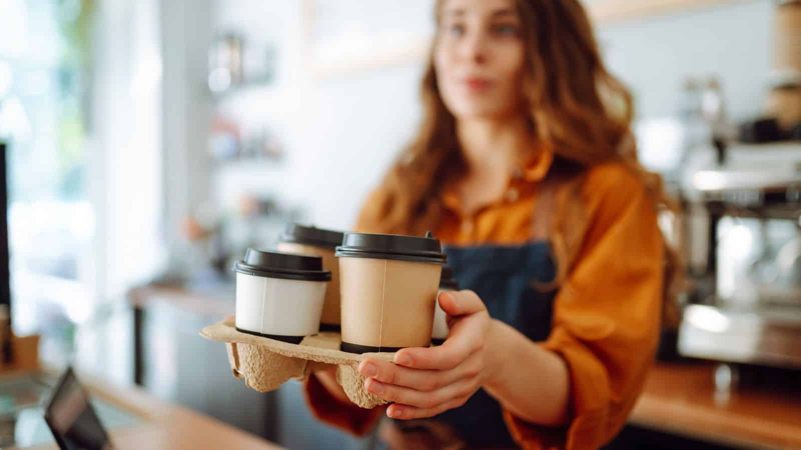 A person wearing an apron holds out a cardboard tray with four lidded coffee cups in a cafe setting. The background shows a coffee machine and blurred interior elements.