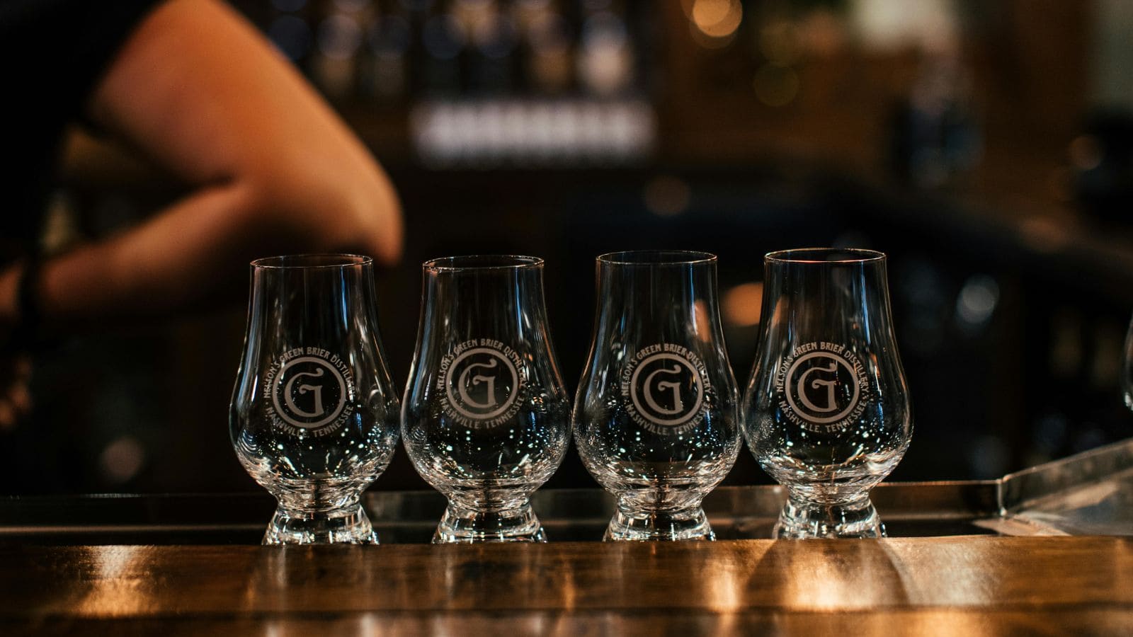 Four empty branded tasting glasses are lined up on a bar counter. A person with crossed arms is partially visible in the background, and the bar area is filled with warm lighting and blurred details.