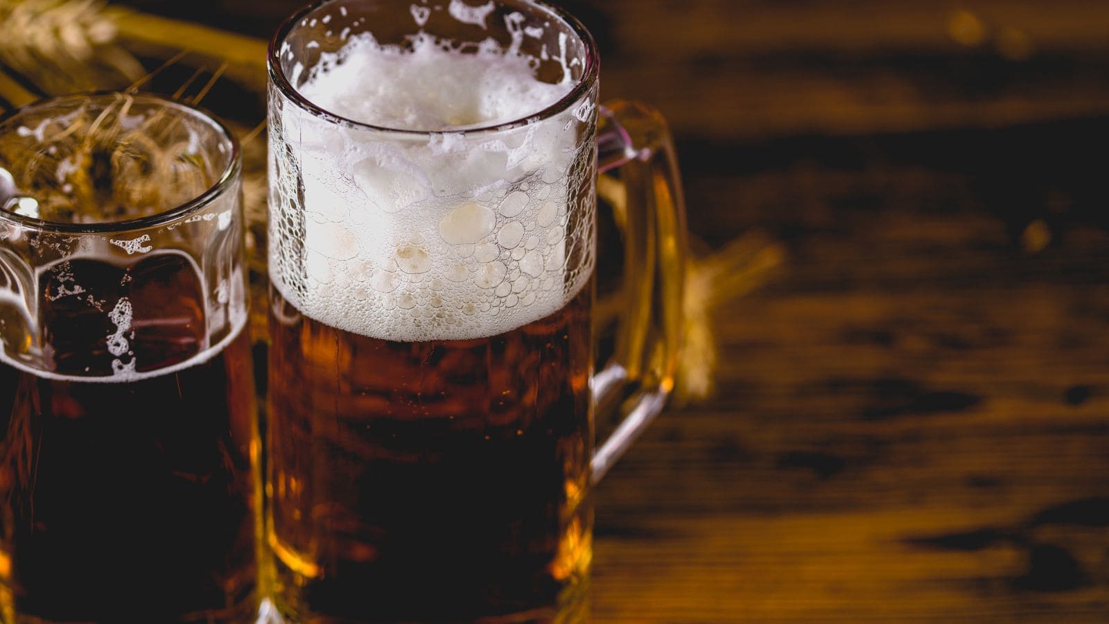 A close-up of two glass mugs filled with dark beer, one with a thick, foamy head. The mugs are on a rustic wooden surface, with wheat stalks in the background.