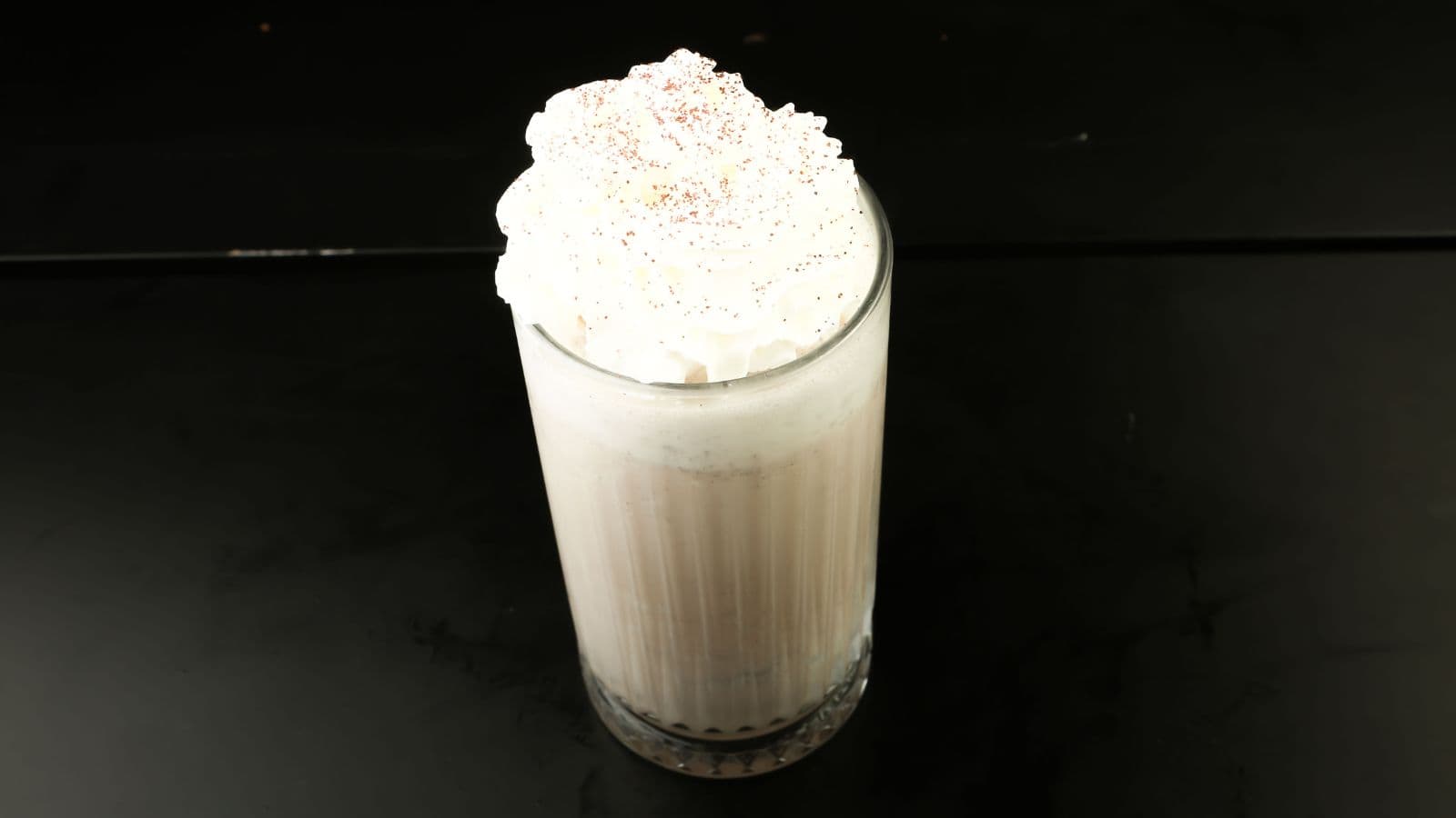 A glass filled with a light beige milkshake topped with a large swirl of whipped cream and a sprinkle of brown powder, set against a plain black background.