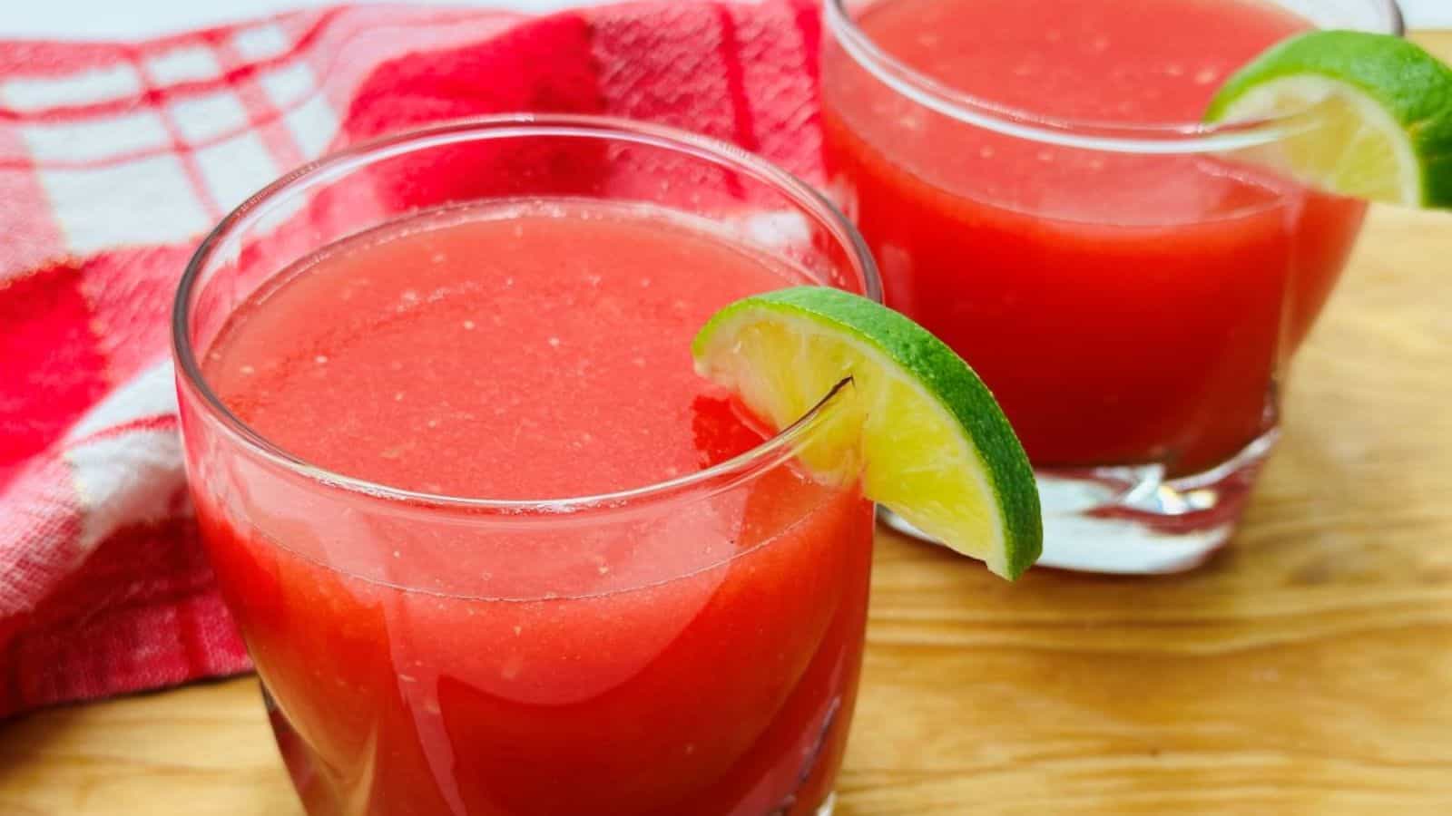 Two glasses of watermelon juice garnished with lime slices are on a wooden surface with a red napkin in the background.