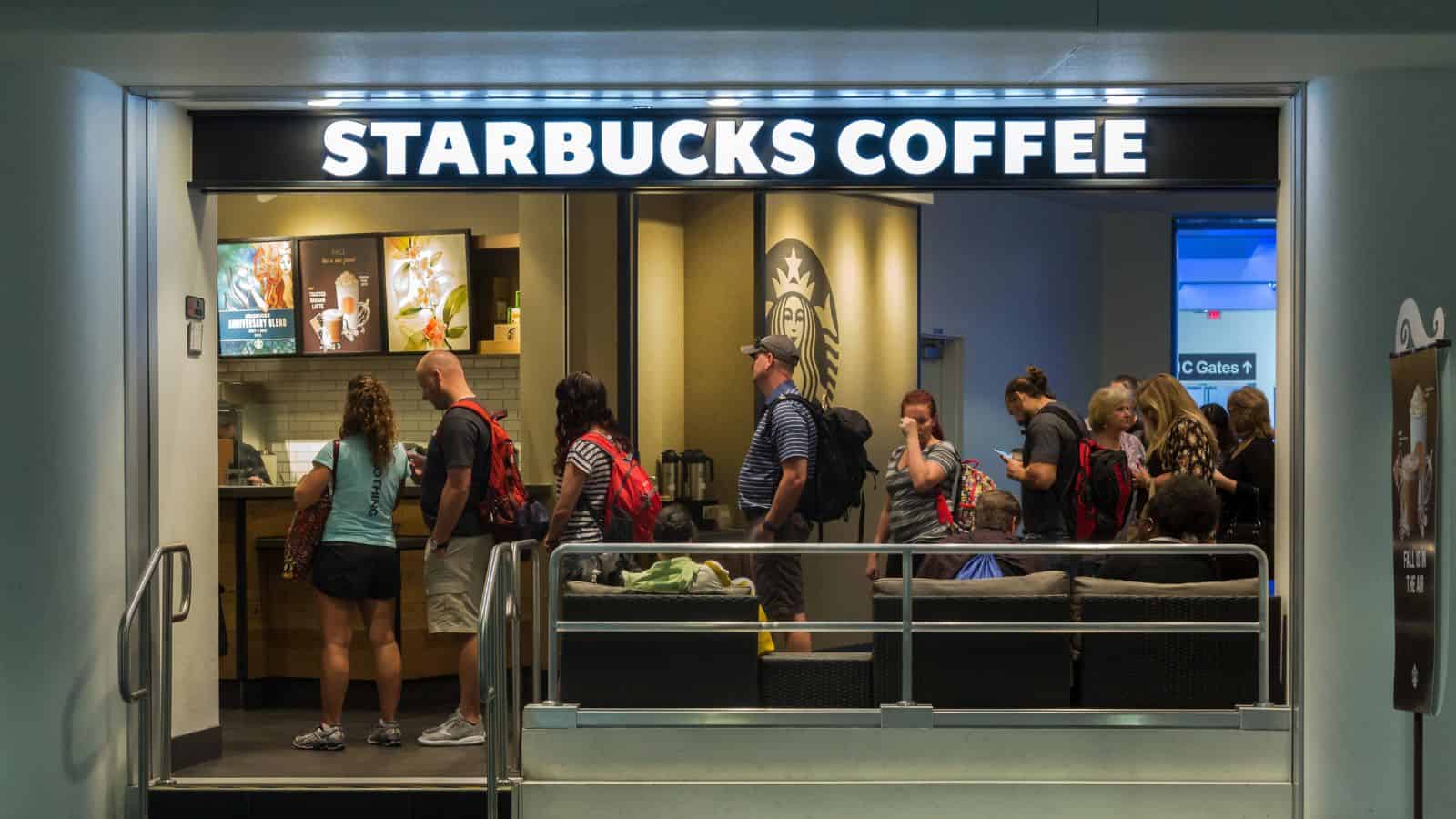 People standing in line inside a Starbucks Coffee shop. The store has a large lighted sign reading "Starbucks Coffee" above the entrance. Some customers are placing orders at the counter, and others are sitting on a couch.