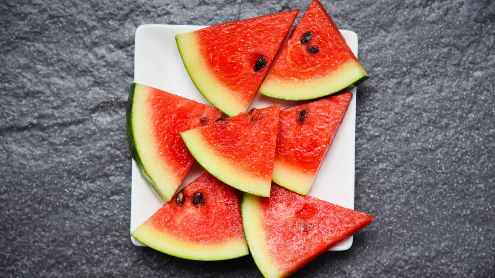 Several triangular slices of watermelon with seeds are arranged on a white rectangular plate, placed on a textured grey surface.