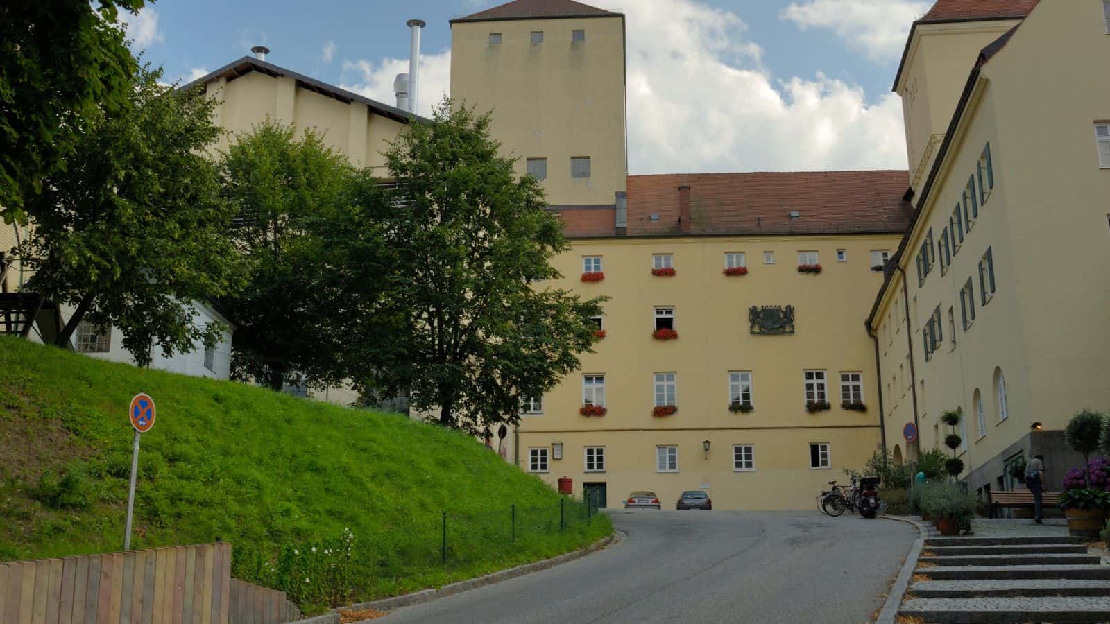 A beige building with a red tiled roof sits on a slight hill. It has multiple windows, some adorned with flower boxes. A narrow road curves around the building, with trees and green grass on the left. Motorcycles and cars are parked nearby.
