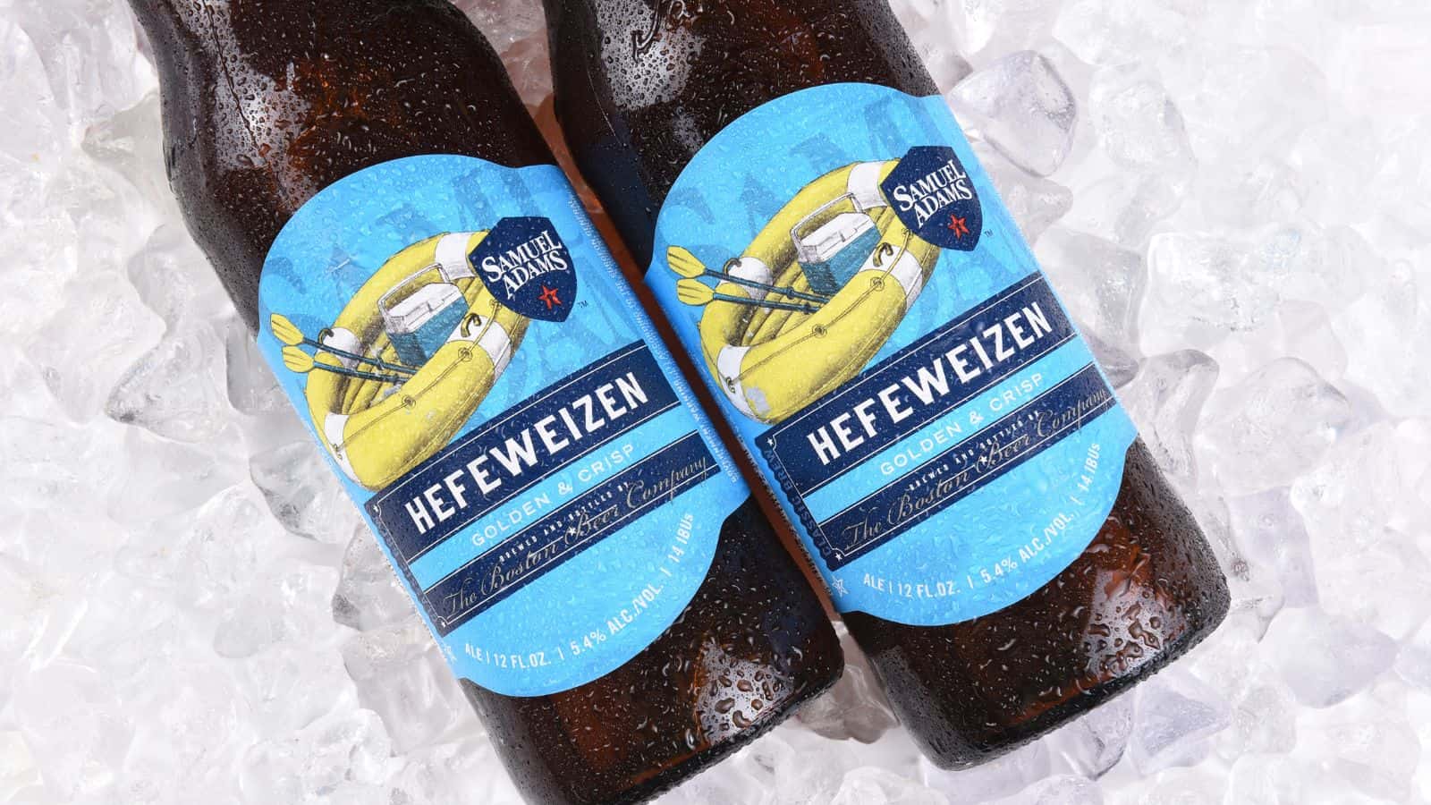 Two bottles of Samuel Adams Hefeweizen lie on crushed ice. The labels are blue with images of wheat, lemons, and a banana, advertising the beer as "Golden & Crisp." Water droplets cover the bottles, suggesting they are cold.