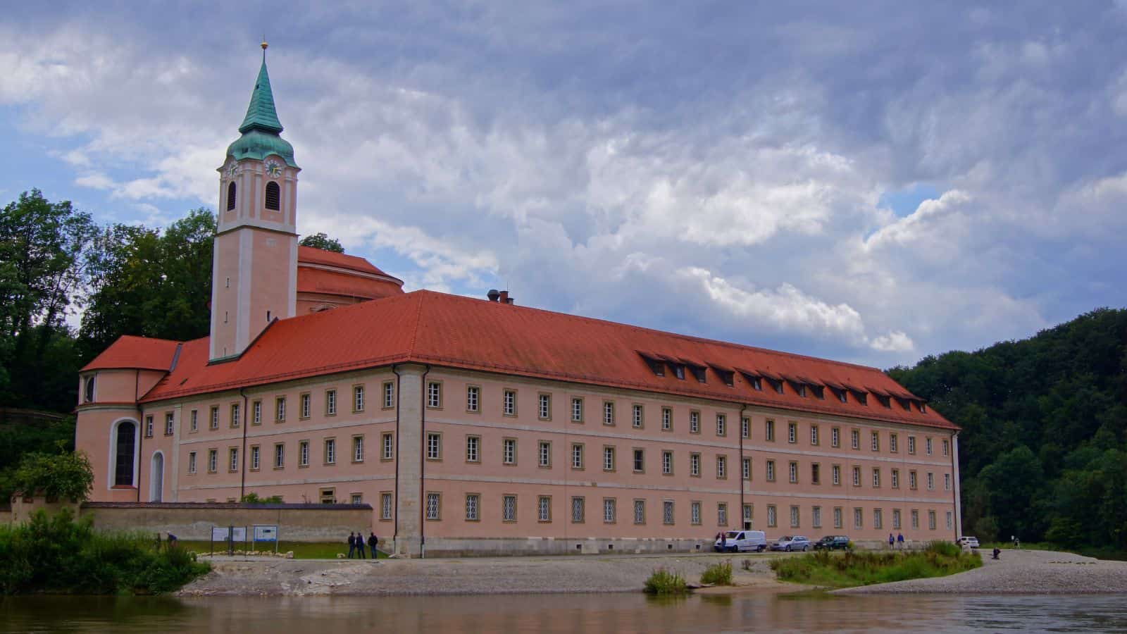 A large, pale pink building with a red-tiled roof and a tall clock tower with a green spire sits by a riverbank. Trees surround the structure, and a cloudy sky is in the background.