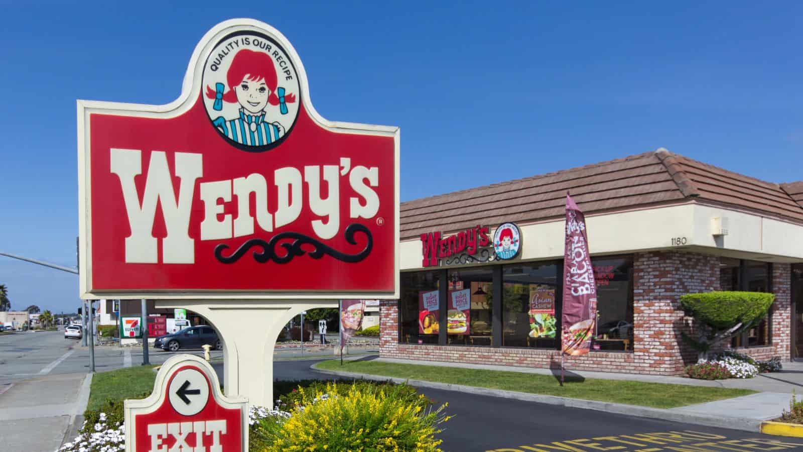 A Wendy’s restaurant with a large red and white Wendy’s sign in the foreground and the restaurant building with windows, signage, and a brown roof in the background on a sunny day.