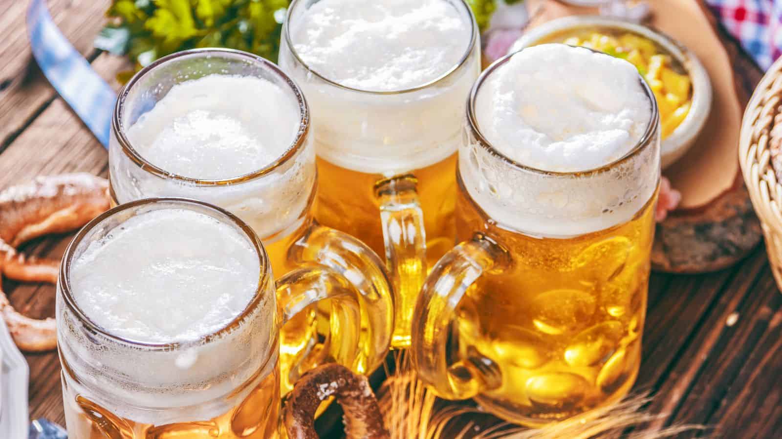 Four glass mugs filled with frothy beer are placed on a wooden table. Surrounding the mugs are wheat stalks and pretzels. In the background, there's a bowl containing a sauerkraut-like dish. Greenery is partially visible.