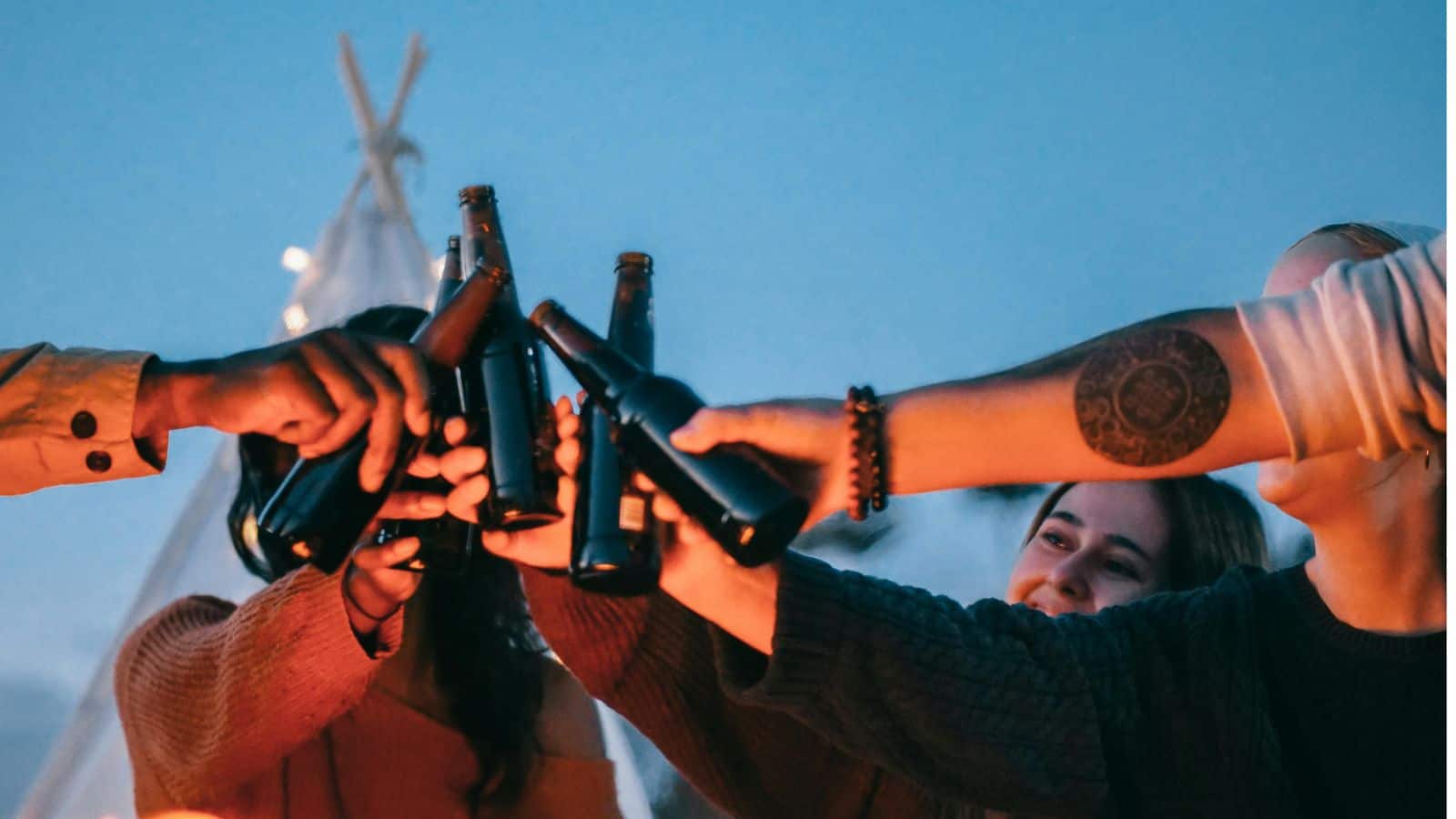 A group of people clink brown glass beer bottles together in a toast outdoors at dusk. Some arms are visible, and one person has a visible tattoo; everyone appears to be smiling and enjoying the moment.