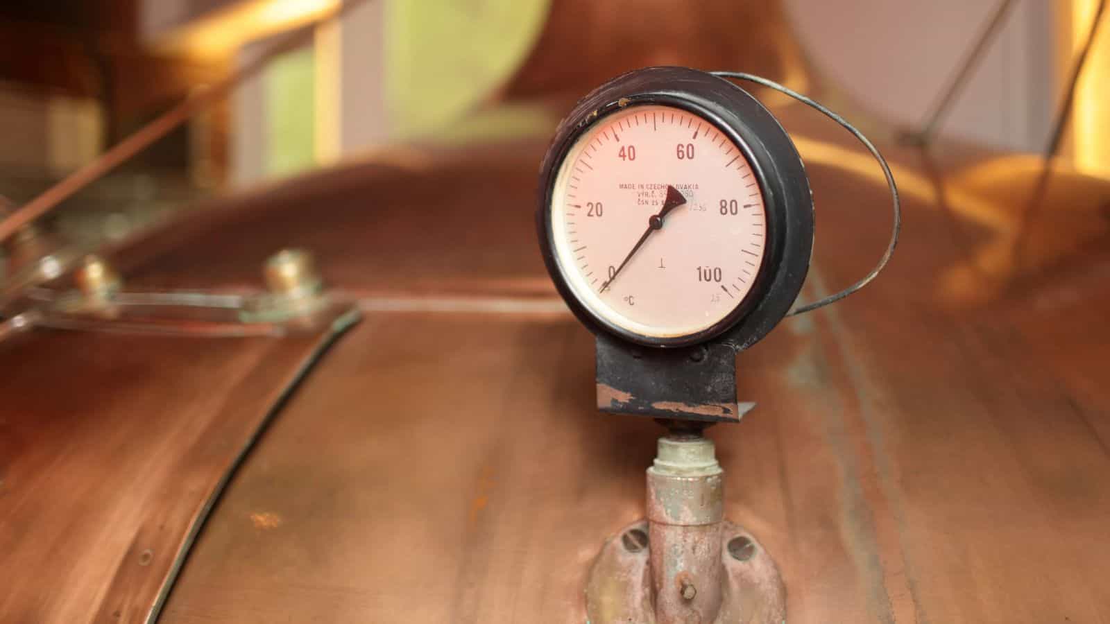 A close-up of a pressure gauge on a copper surface, showing a needle pointing to around 60. The gauge has black markings from 0 to 100 and a metal casing. The background is slightly blurred and features copper and metal elements.