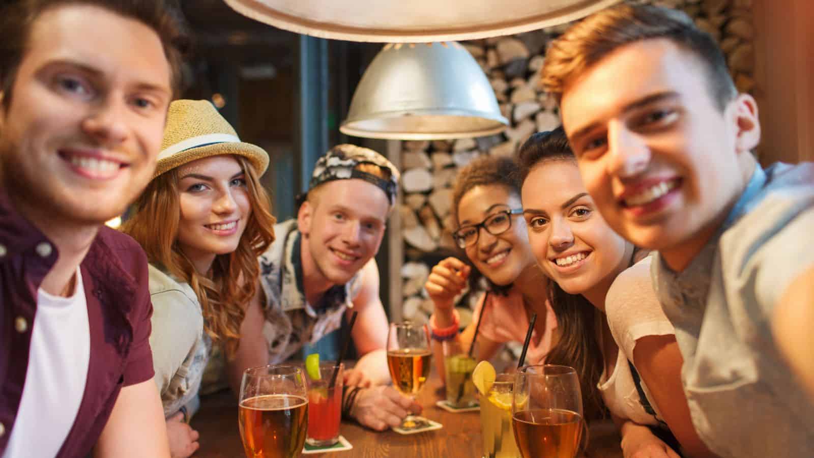 Six young adults sit closely together at a wooden table in a bar or restaurant, smiling at the camera. There are several glasses of drinks with straws and garnishes in front of them. Warm lighting hangs overhead.
