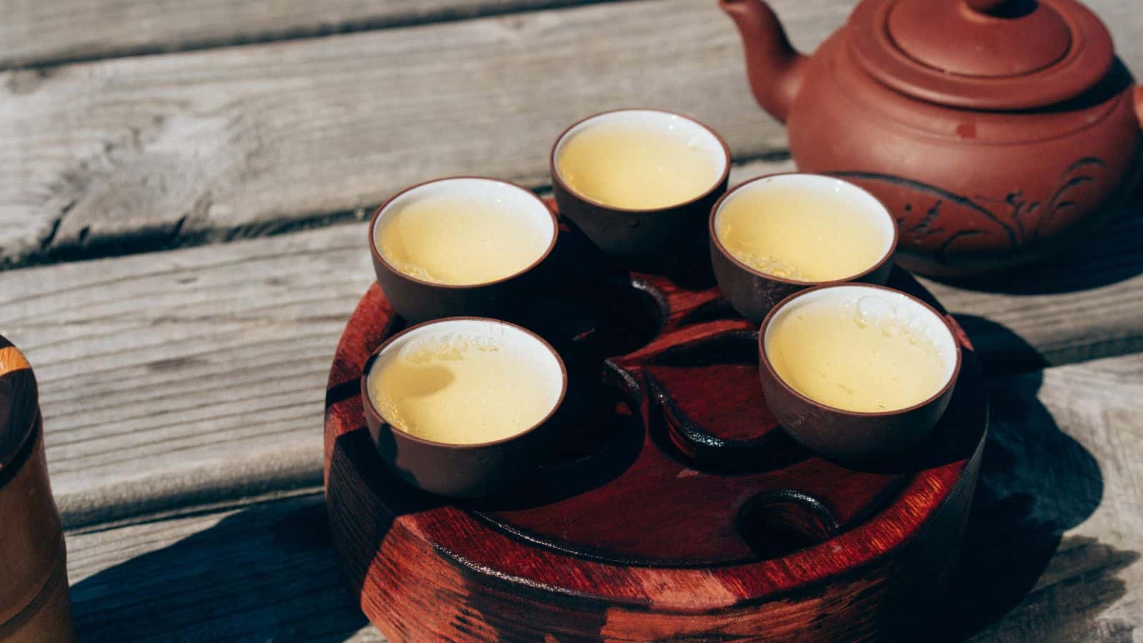 Five small cups of tea sit on a round wooden tray with a carved design, placed on a weathered wooden surface. A brown teapot with decorative patterns is nearby. The tea appears light and steaming.