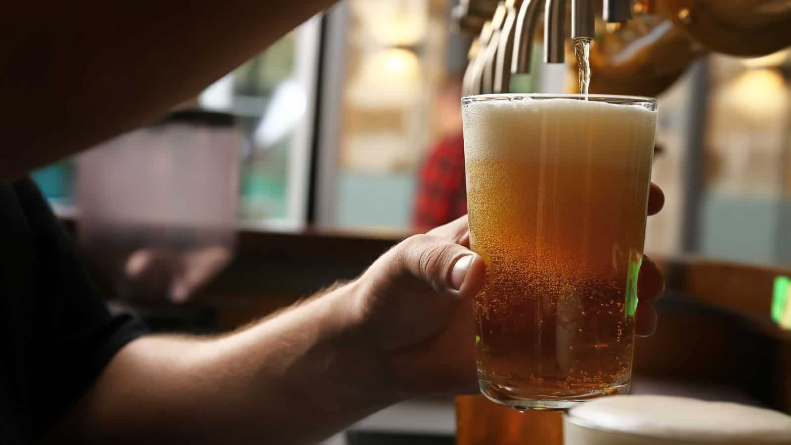 A person holds a clear glass as beer with a foamy head is poured from a tap in a bar or pub setting, with a blurred background.