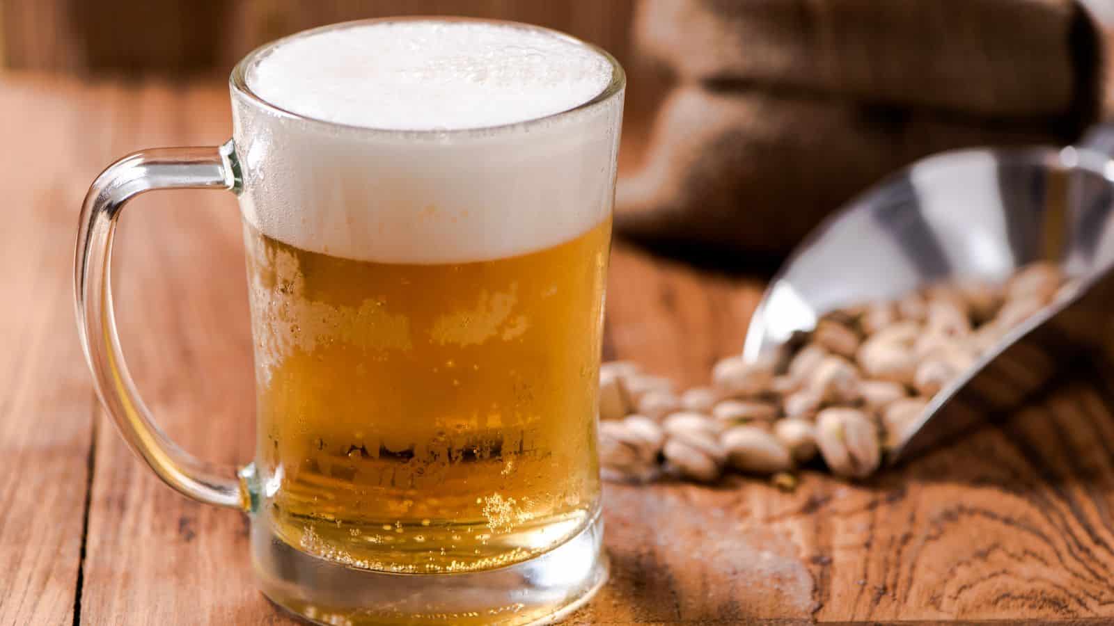 A glass mug of beer with a foamy head sits on a wooden table. Beside it, there is a metal scoop filled with pistachios. A sack partially visible in the background adds a rustic touch.