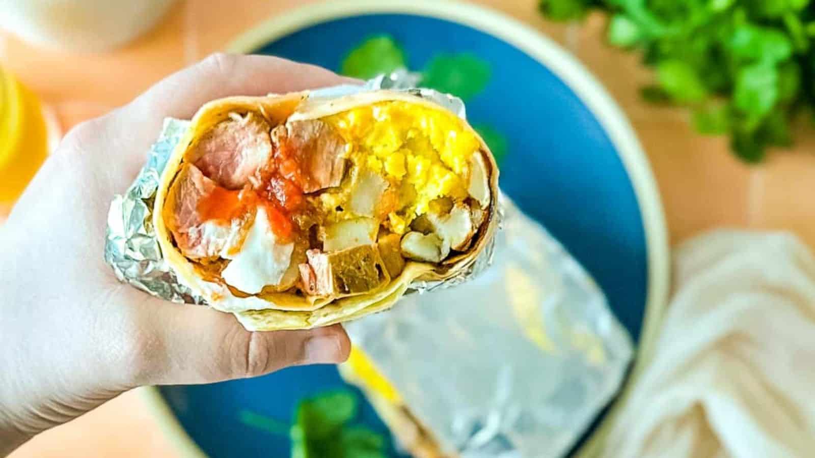 Half of a carne asada breakfast burrito is held over a blue plate surrounded by limes, orange juice, coffee, and a white linen.