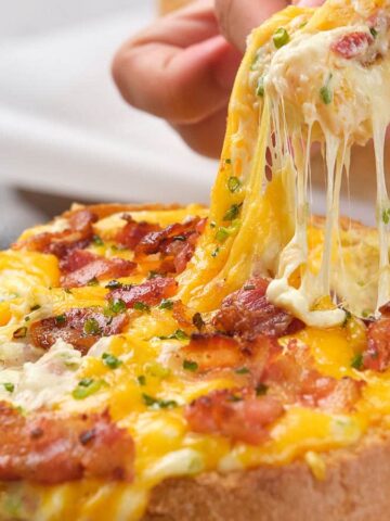 A hand pulling a piece of bread filled with creamy, melted cheese and bacon, creating a stringy effect. The bread bowl is surrounded by pieces of crust. Parsley is sprinkled on top as garnish.