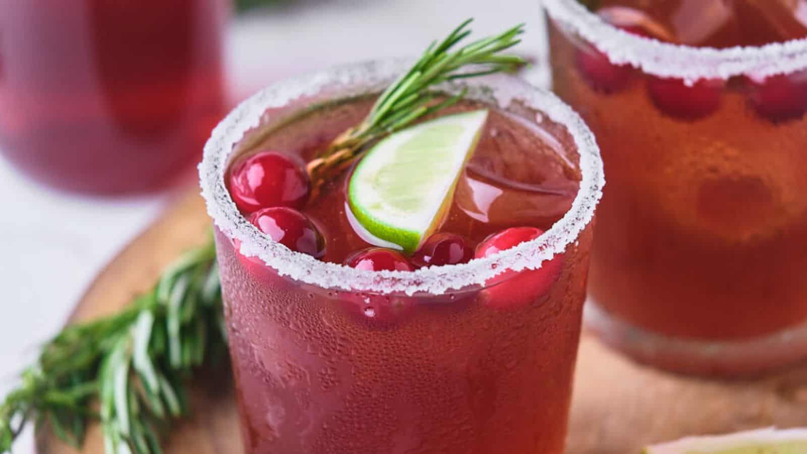 Two glasses of a red cocktail garnished with cranberries, a lime wedge, and a sprig of rosemary.
