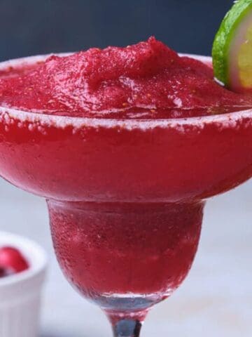 A frozen red drink in a margarita glass with a sugar rim is garnished with a lime slice. A hand is placing the lime on the glass. A bowl of cherries is visible in the background.