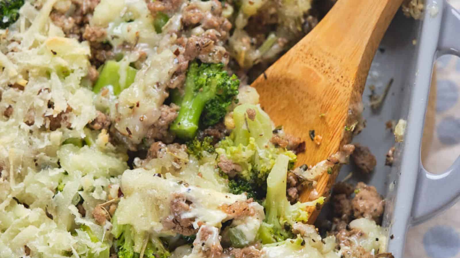 A close-up of a cheesy ground beef and broccoli casserole in a baking dish. The dish features visible pieces of broccoli, ground beef, and melted cheese. A wooden spoon is partially submerged in the casserole.