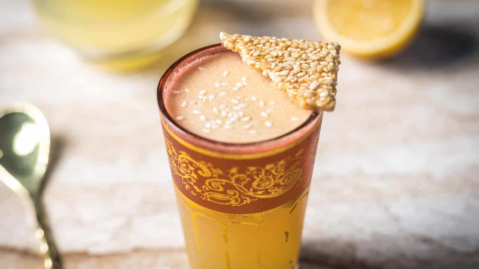 A glass of frothy yellow beverage garnished with sesame seeds and topped with a triangular piece of sesame brittle. A gold spoon, a lemon half, and another glass are in the blurred background.